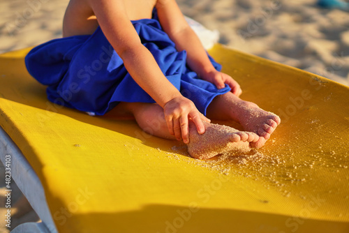 A small child on a sunbed by the sea. A girl in a towel is warming up after bathing. Vacation with a child. The little man's arms and legs are tanning. Sun protection cream.