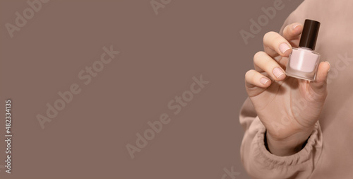 Female hand with beige nail design. Glossy beige fingernails manicure. Woman hand hold beige nail polish bottle on brown background photo