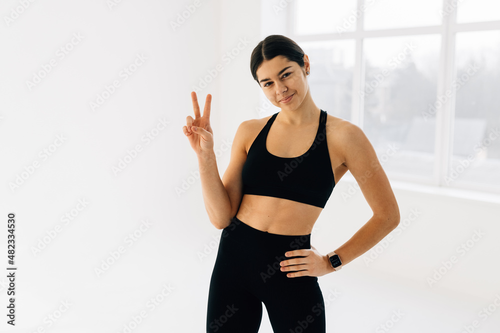 Fit and slim fitness woman showing peace sign and winking, workout in gym, doing sports exercises in activewear, white background Fit and slim fitness woman showing peace sign and winking, workout in gym, doing sports exercises in activewear, white background