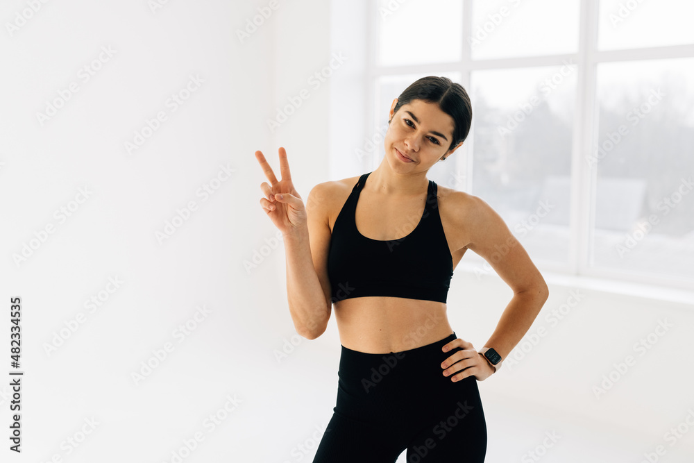 Fit and slim fitness woman showing peace sign and winking, workout in gym, doing sports exercises in activewear, white background Fit and slim fitness woman showing peace sign and winking, workout in gym, doing sports exercises in activewear, white background