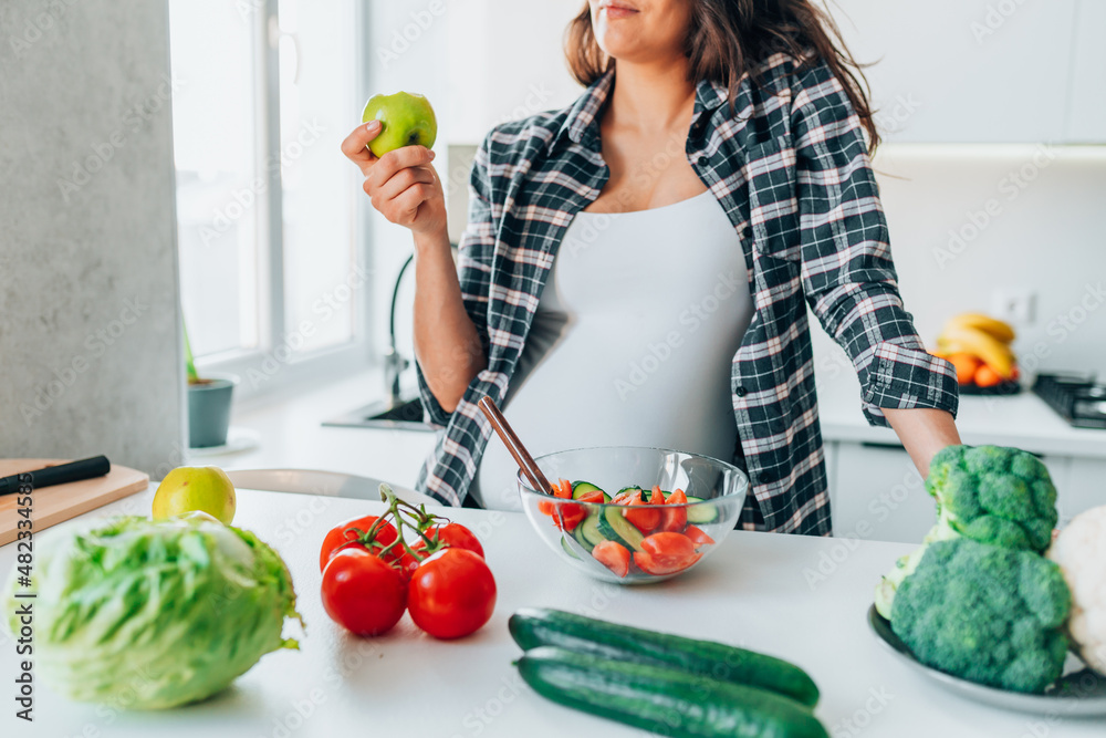 Eat healthy during pregnancy. Cropped image of young pregnant woman preparing fresh vegetable salad, standing behind table in kitchen at home, selective focus. Expectant mother eating green apple Eat healthy during pregnancy. Cropped image of young pregnant woman preparing fresh vegetable salad, standing behind table in kitchen at home, selective focus. Expectant mother eating green apple
