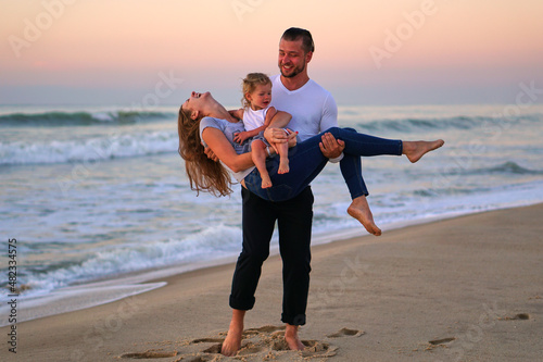 Happy family on the beach. A man holds a woman and a girl in his arms. Parents and daughter have fun on the beach. Traveling with children. Be together on the beach. High quality photo