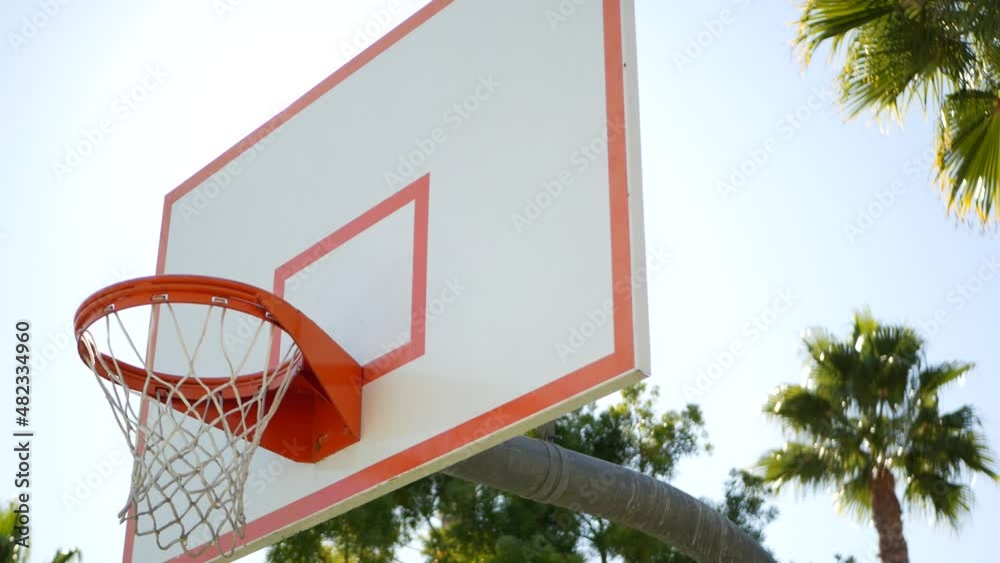 Basketball court outdoors, orange hoop, net and backboard for basket ball game outside. Recreational sport equipment on streetball field or playground. Blue sky and beach palm trees, California USA. Basketball court outdoors, orange hoop, net and backboard for basket ball game outside. Recreational sport equipment on streetball field or playground. Blue sky and beach palm trees, California USA.
