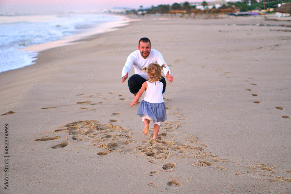 A little girl runs to her dad on the sand by the sea. The child is happy to travel with his parents. A man meets a girl beach running towards her father. open arms father on beach at the sunny day A little girl runs to her dad on the sand by the sea. The child is happy to travel with his parents. A man meets a girl beach running towards her father. open arms father on beach at the sunny day