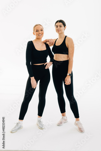 Fototapeta Naklejka Na Ścianę i Meble -  Two concentrated women dressed in sports wear bonding to each other while standing indoor against white background. People, exercising and sport concept