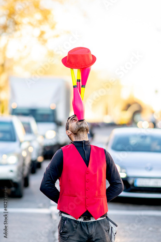 Performer balancing juggling pins and hat on nose in front of cars photo