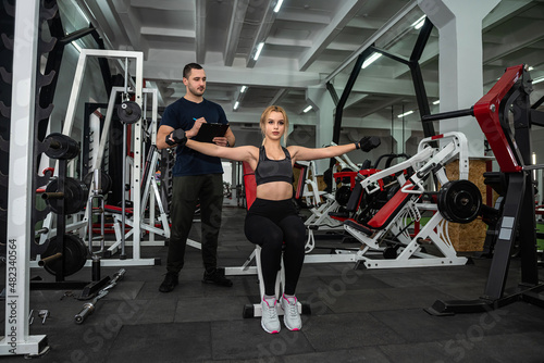  trainer with athletic body helps girl client to perform proper exercises on the simulator.