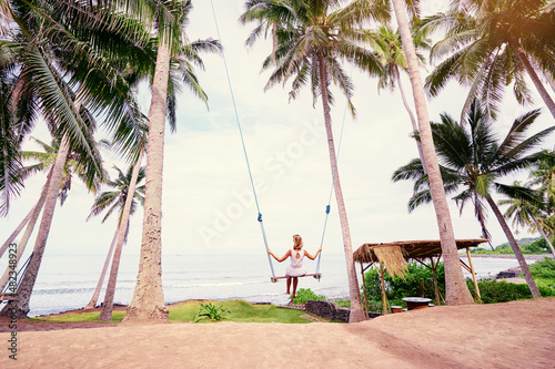 Vacation concept. Happy young woman in white dress and hat swinging at palm grove enjoying sea view. photo