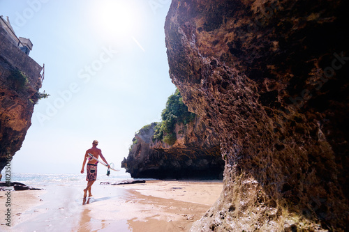 Hobby and vacation. Young man with surfboard on beautiful beach with high rocks. Uluwatu spot, Bali island, Indonesia.
