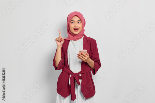 Excited beautiful Asian woman in casual shirt and hijab holding mobile phone and pointing finger up, creating genius solutions isolated over white background. People religious lifestyle concept