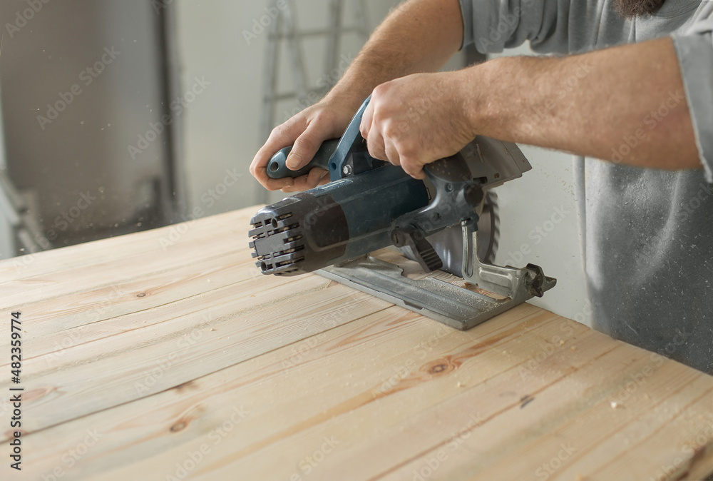 a man with a beard in a helmet saws a wooden board circular hand saw. a man with a beard in a helmet saws a wooden board circular hand saw.