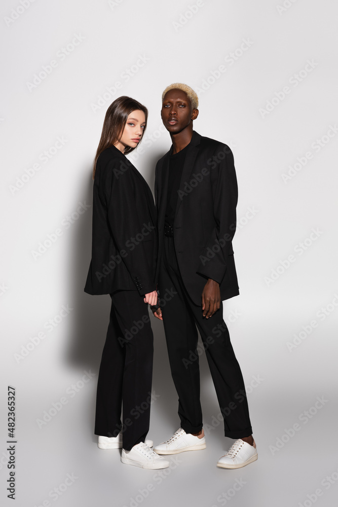 full length view of trendy interracial couple in black suits and white sneakers on grey with shadow.