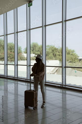 young girl is standing near window at the airport before departure, model dressed in white pants, pink jacket and hat, pink suitcase in hands © Zhenia