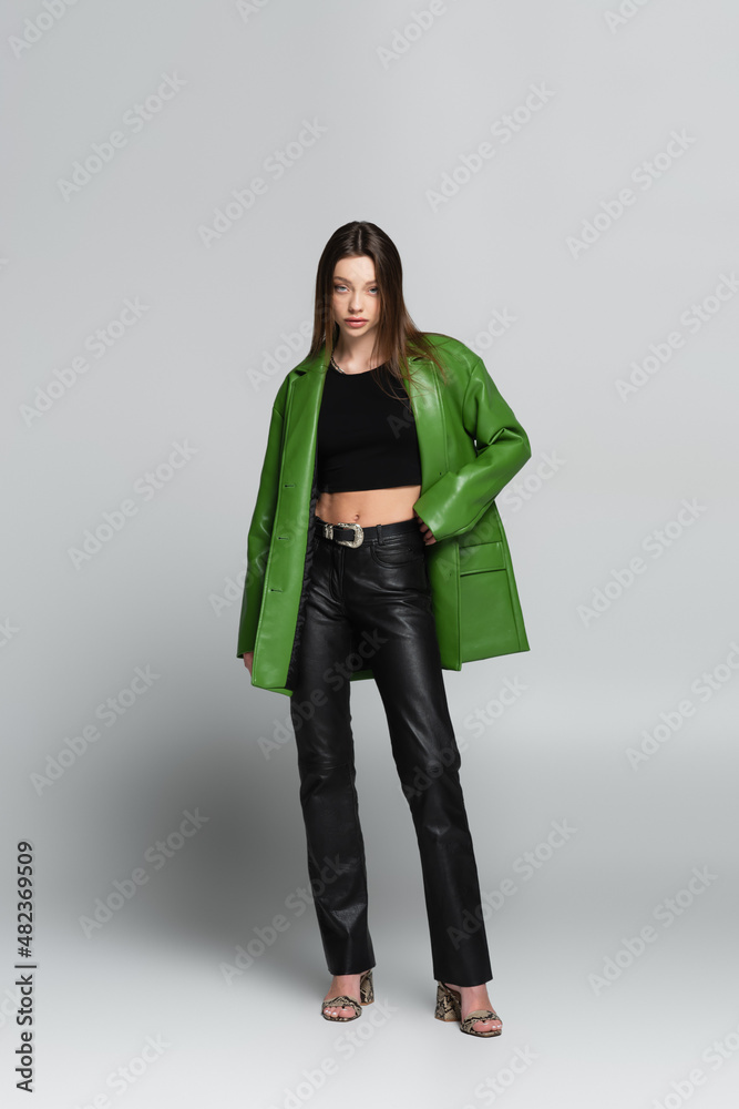 front view of brunette woman in black trousers and green jacket standing with hand on waist on grey. front view of brunette woman in black trousers and green jacket standing with hand on waist on grey.