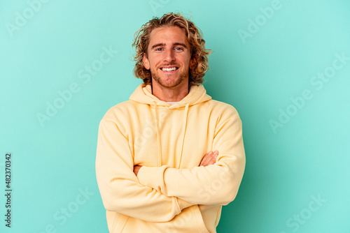 Young caucasian man isolated on blue background who feels confident, crossing arms with determination.