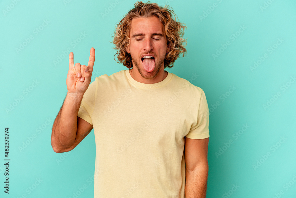 Young caucasian man isolated on blue background showing rock gesture with fingers Young caucasian man isolated on blue background showing rock gesture with fingers