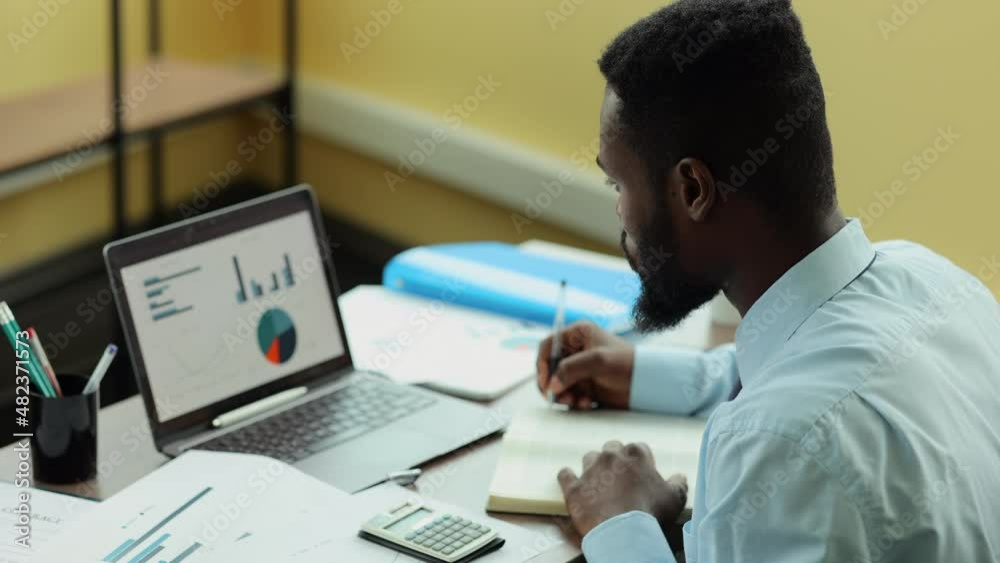 Businessman or analyst working on online project and writing while sitting at table in office spbi. Closeup view of American African man looks at charts on computer screen and writes in notebook, does