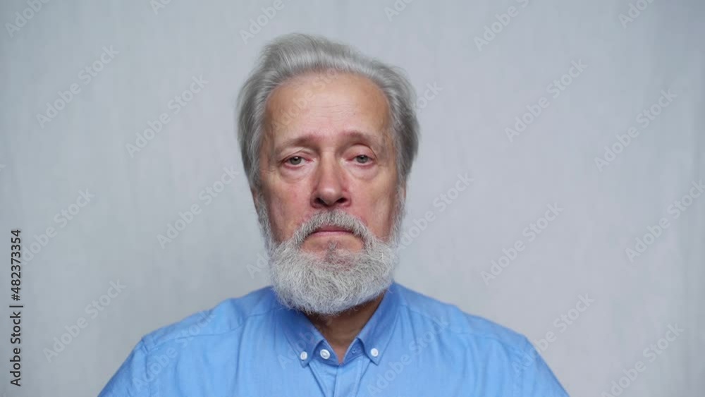 Close-up portrait of serious gray-haired mature man unemotionally looking at camera standing on white isolated background. Front view of bearded old aged gentleman posing in studio, slow motion. Close-up portrait of serious gray-haired mature man unemotionally looking at camera standing on white isolated background. Front view of bearded old aged gentleman posing in studio, slow motion.