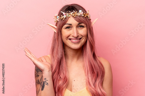 Young caucasian elf woman isolated on pink background showing a copy space on a palm and holding another hand on waist.