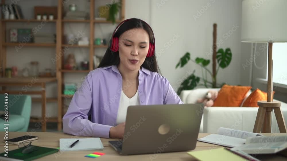Female student watching online webinar and studying in front of laptop at table in apartment spbd. Front view of beautiful Asian woman looks at computer screen and speaks with smile, watches lecture Female student watching online webinar and studying in front of laptop at table in apartment spbd. Front view of beautiful Asian woman looks at computer screen and speaks with smile, watches lecture