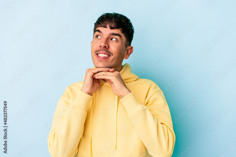 Young mixed race man isolated on blue background keeps hands under chin, is looking happily aside. Young mixed race man isolated on blue background keeps hands under chin, is looking happily aside.