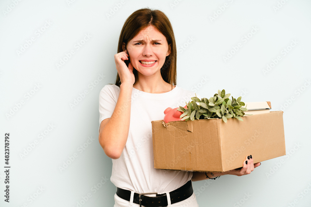 Young English woman making a move while picking up a box full of things isolated on blue background covering ears with hands. Young English woman making a move while picking up a box full of things isolated on blue background covering ears with hands.