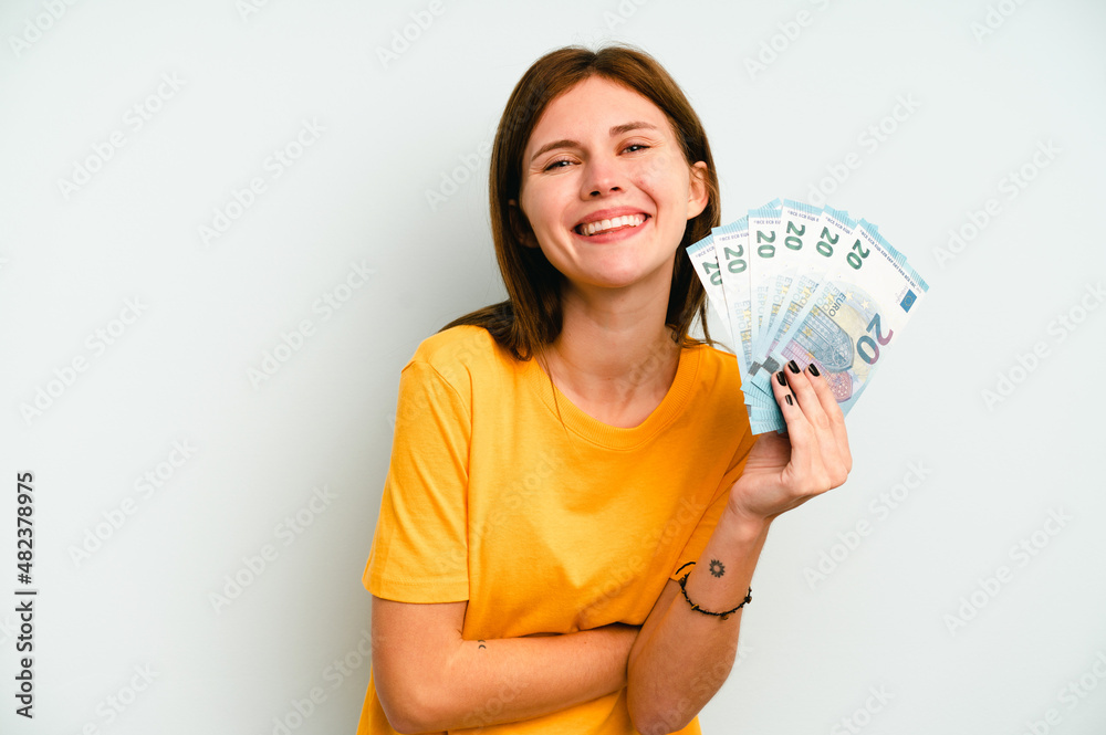 Young English woman holding banknotes isolated on blue background laughing and having fun. Young English woman holding banknotes isolated on blue background laughing and having fun.