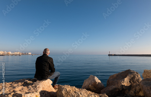 Rear view of adult man sitting on rock with lighthouse and city in background. Almeria, Spain