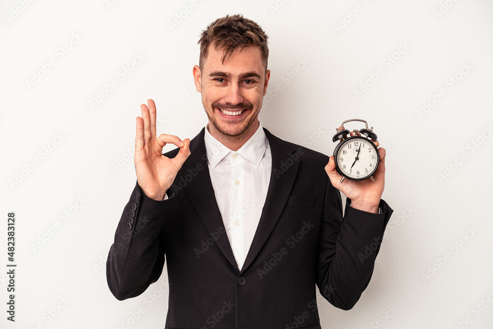Young caucasian business man holding alarm clock isolated on white background cheerful and confident showing ok gesture. Young caucasian business man holding alarm clock isolated on white background cheerful and confident showing ok gesture.