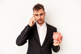 Young caucasian business man holding piggy bank isolated on white background pointing temple with finger, thinking, focused on a task.