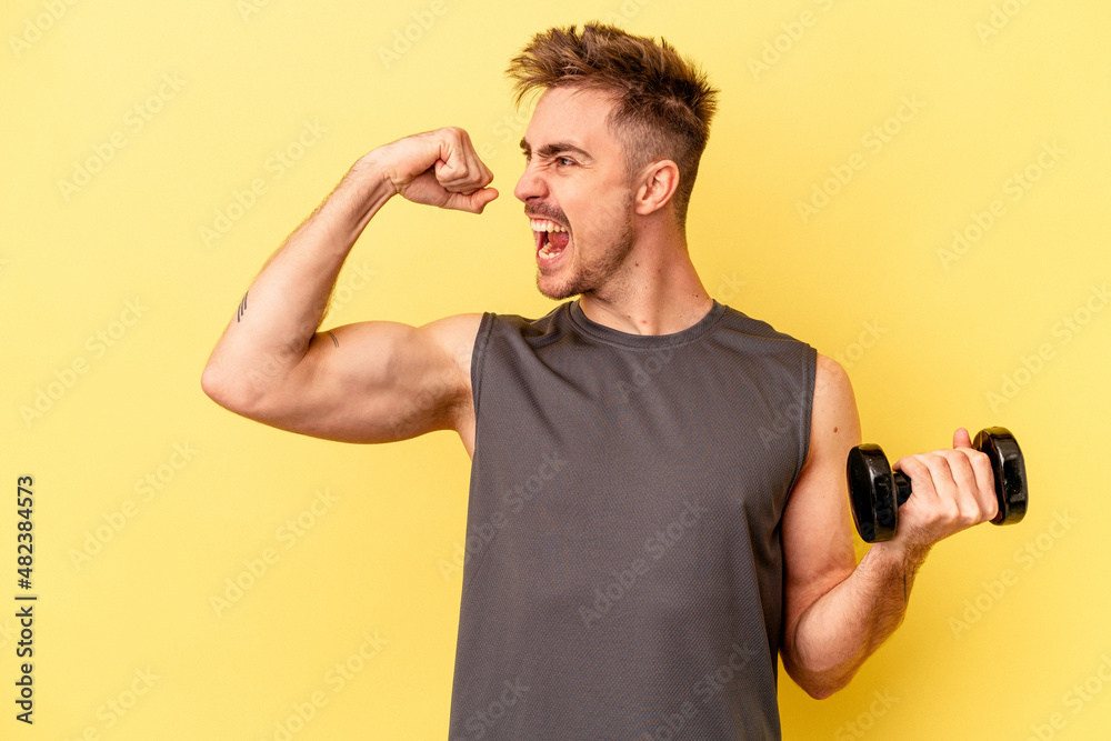 Young sport man holding a dumbbell isolated on yellow background raising fist after a victory, winner concept. Young sport man holding a dumbbell isolated on yellow background raising fist after a victory, winner concept.