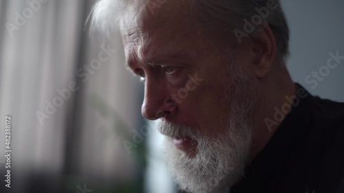 Close-up headshot of gray-haired upset depressed mature adult man crying alone at home thinking of loneliness grief. Closeup face of sad senior male in tears worried about health problems, slow motion