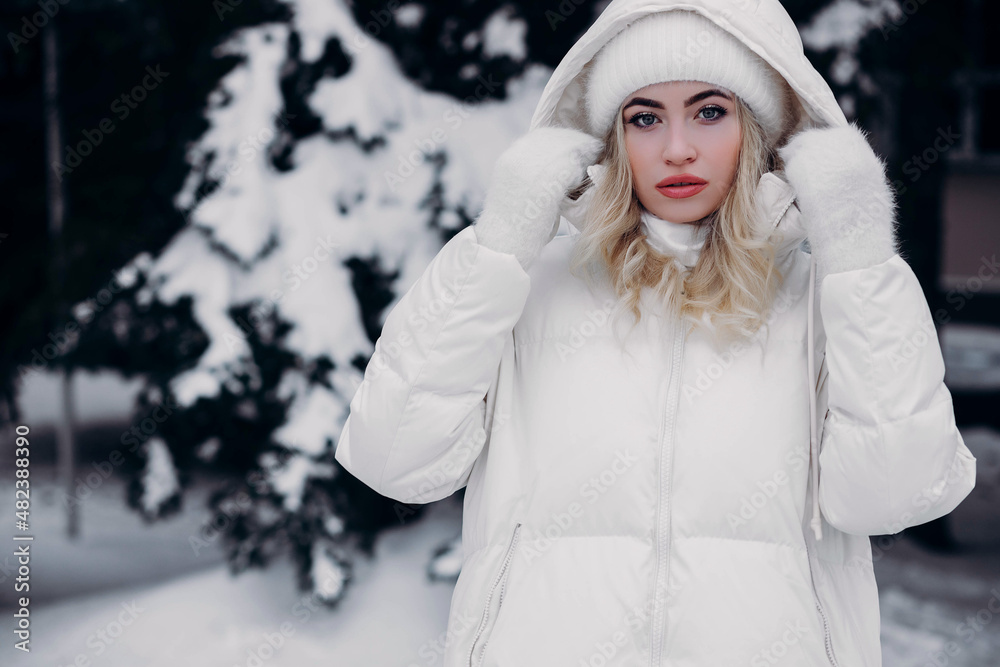 Portrait of a young woman in winter, outdoors. The girl holds on to the hood of a white jacket and smiles. A very cold, snowy winter day. Large winter portrait. Portrait of a young woman in winter, outdoors. The girl holds on to the hood of a white jacket and smiles. A very cold, snowy winter day. Large winter portrait.