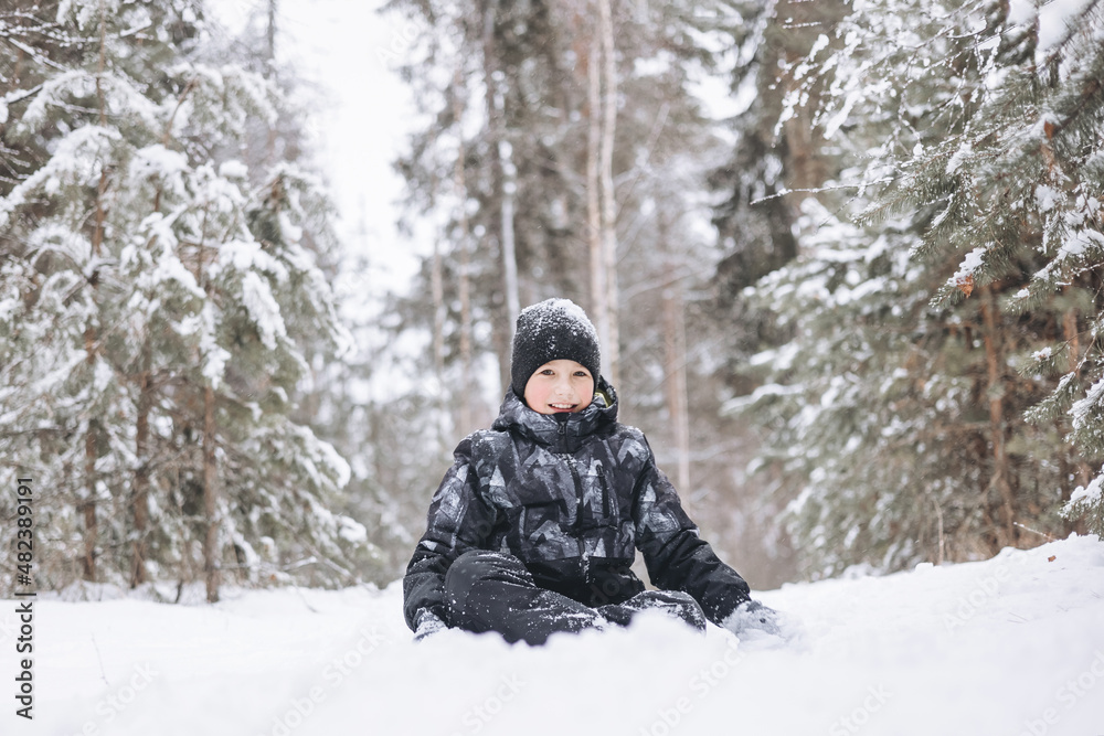Happy teenager boy sitting on snow in winter forest. Child having fun outdoors. Joyful adolescent playing in snow at snowfall. Laughing smiling kid walking in winter park in cold weather