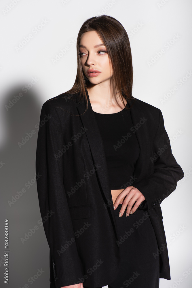 Fototapeta premium stylish woman in black clothes standing with hand on waist and looking away on grey with shadow.