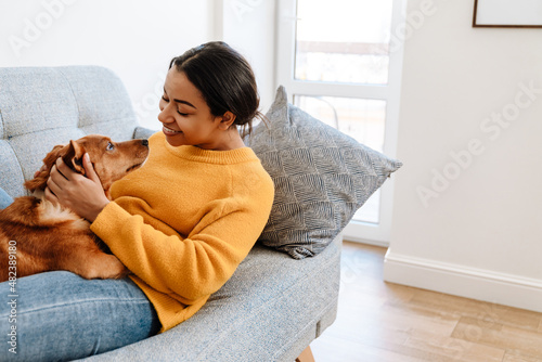 Young hispanic woman smiling and petting her dog while resting on couch
