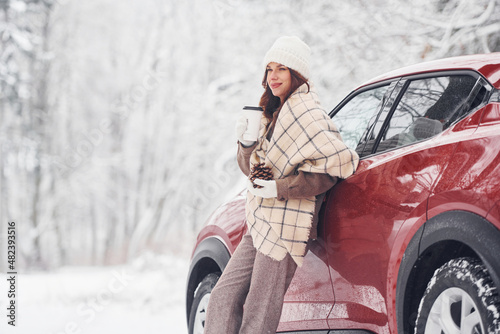 With cup of drink. Beautiful young woman is outdoors near her red automobile at winter time