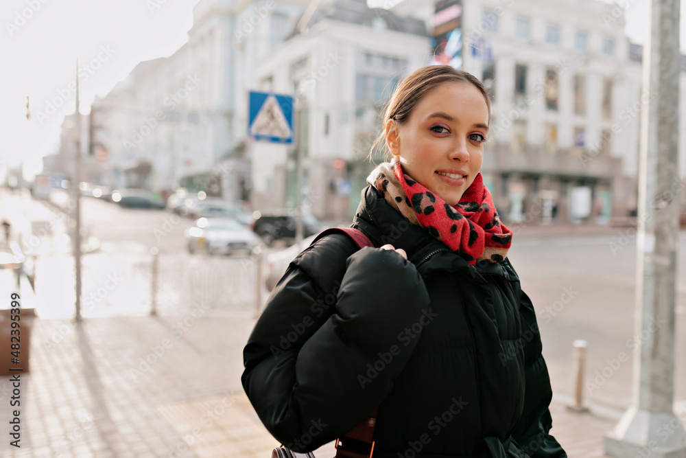 Glamour stylish girl with bright make up wearing black jacket and colorful scarf walking in the city in sunshine and smiling at camera. Happy true emotions, good warm day