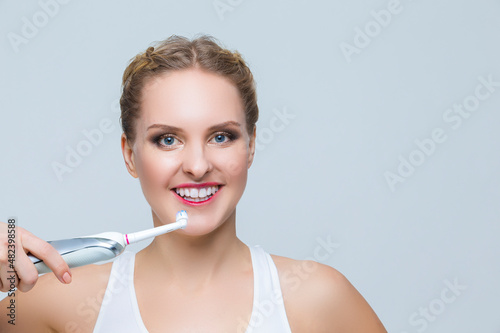 Dental Healthcare. Smiling Caucasian Blond Woman in Bra Cleaning Teeth with Electric  Toothbrush Over White