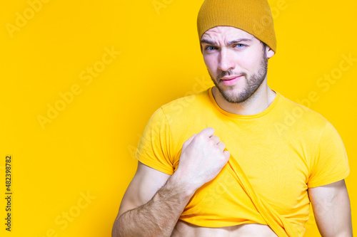Serious Caucasian Handsome Man In Yellow Tshirt Posing in Warm Hat Over Yellow Background While Pulling Shirt Off