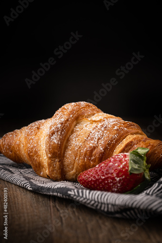 breakfast croissant with powdered sugar and strawberry dark background with black and white napkin.
