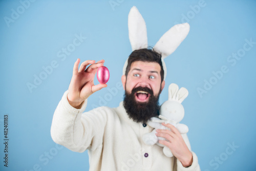 Hipster cute bunny long ears blue background. Easter bunny. Having fun. My precious. Funny bunny with beard and mustache hold pink egg. Easter symbol concept. Bearded man wear silly bunny ears photo