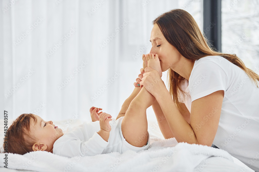 On the white bed. Mother with her little daughter is indoors at home together On the white bed. Mother with her little daughter is indoors at home together