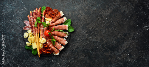 Italian ingredients: ham, prosciutto, salami, parmesan, olives, bread sticks. On a black stone background.