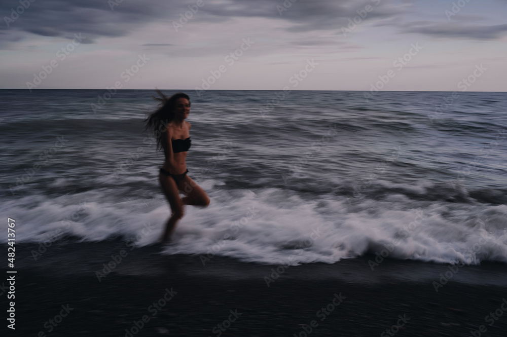 happy young girl in a swimsuit runs on sea waves on beach on a summer evening. Photo with a blur from a long exposure to give an authentic atmosphere happy young girl in a swimsuit runs on sea waves on beach on a summer evening. Photo with a blur from a long exposure to give an authentic atmosphere