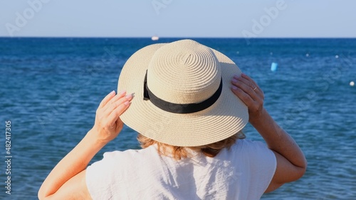 Back view of a woman in a straw hat and white dress looking at the blue sea, close up. The concept of relaxation, rest, sunscreen