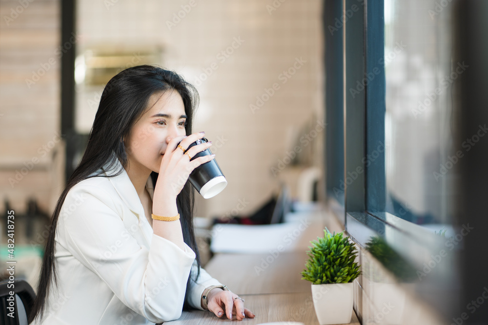 Portrait of a young Asian woman sipping coffee and looking thoughtfully out the window of a coffee shop Portrait of a young Asian woman sipping coffee and looking thoughtfully out the window of a coffee shop