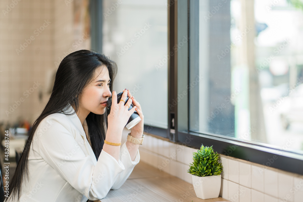Portrait of a young Asian woman sipping coffee and looking thoughtfully out the window of a coffee shop Portrait of a young Asian woman sipping coffee and looking thoughtfully out the window of a coffee shop