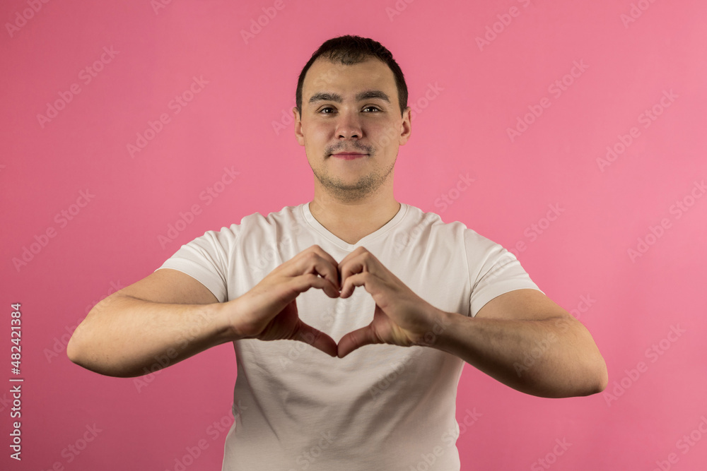 A young handsome man in a white T-shirt on an isolated pink background makes a heart shape with his hands. Concept: love, Valentine's Day, romance A young handsome man in a white T-shirt on an isolated pink background makes a heart shape with his hands. Concept: love, Valentine's Day, romance