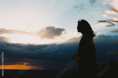 The silhouette of a young woman stands on the rocks at the top of the mountain and looks at the sunset.